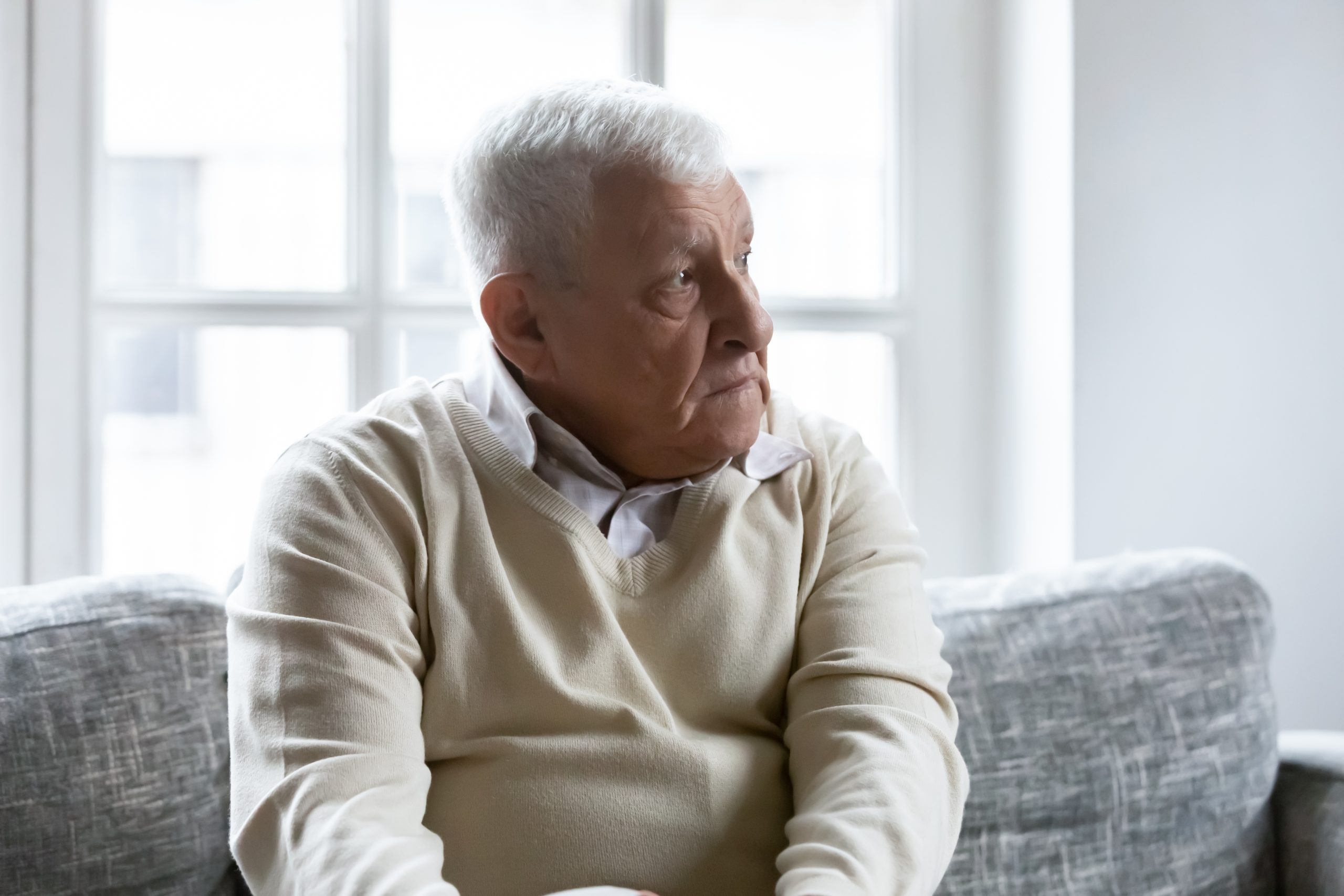 Lost in memories. Sad thoughtful retired grey haired man sits on soft couch at home.   